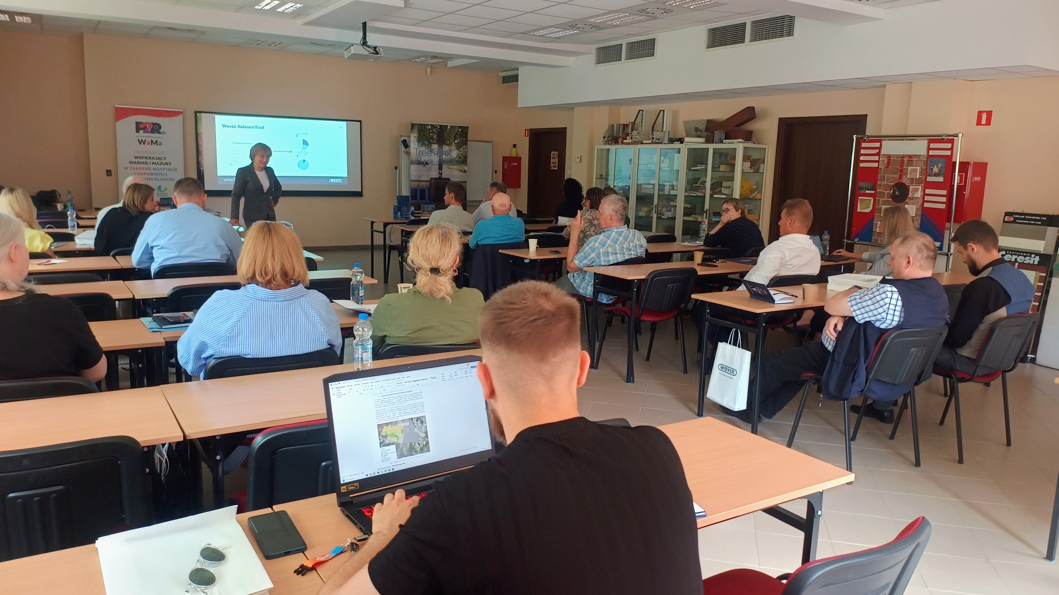 Widok na salę, w której odbywają się zajęcia. Widać uczestników siedzących tyłem do obiektywu, zwróconych w stronę prowadzącego, który znajduje się na przodzie sali.